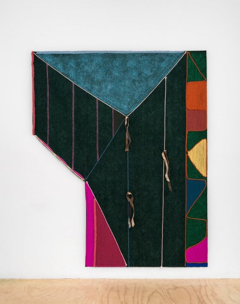 A flat, asymmetrical artwork with a geometric design in black, green, magenta, orange, red and yellow by Teresa Baker hangs on a white wall. The work is made with parfleche, acrylic, artificial sinew, and yarn on astroturf.