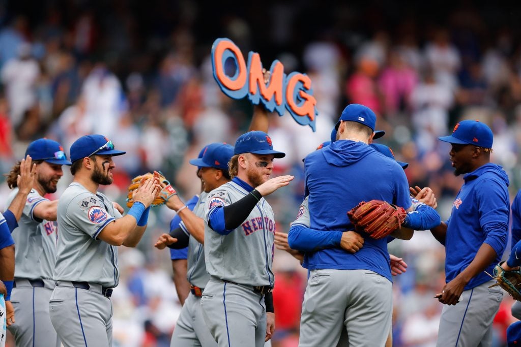 Baseball players hugging and high-fiving each other on the field, one of them brandishing a blue and orange sign that reads "OMG"