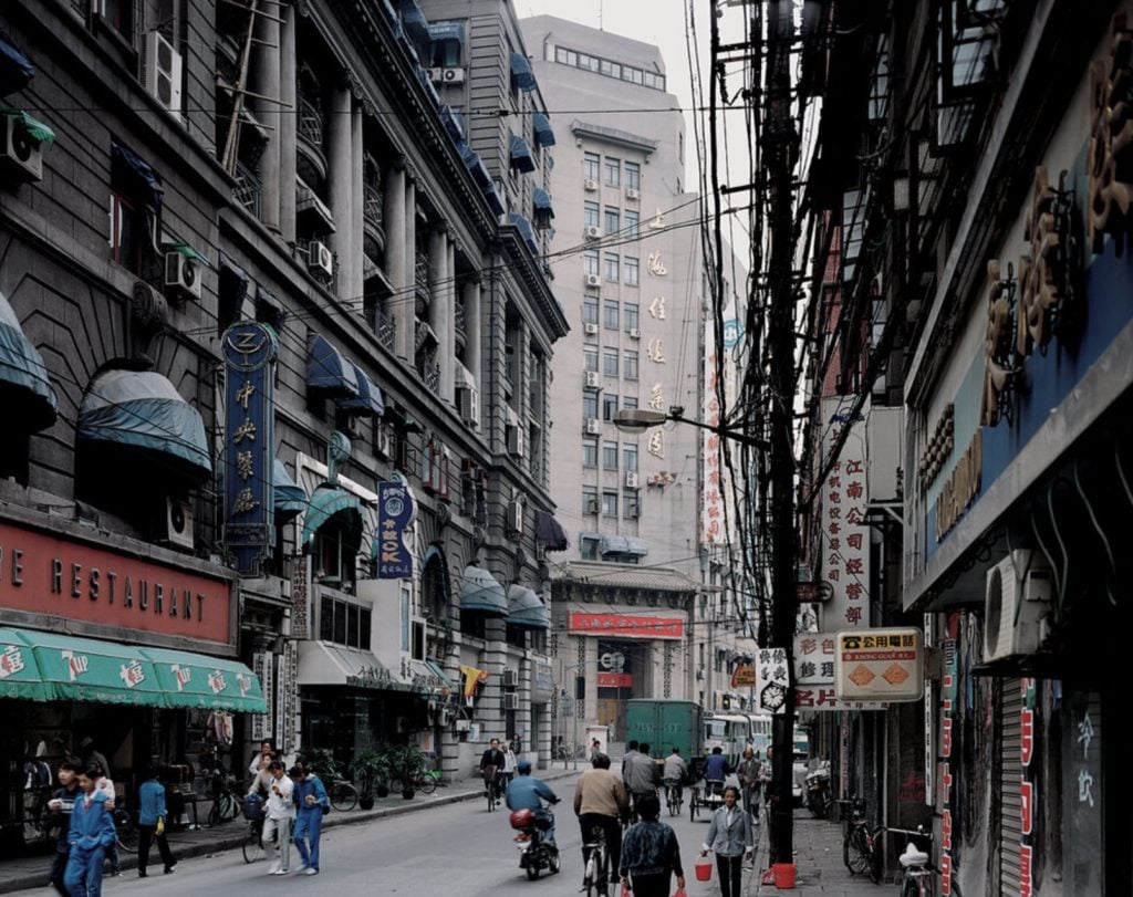 Chromogenic vintage image of a commercial street in Shanghai lined with shops and people walking or riding by on bicycles.