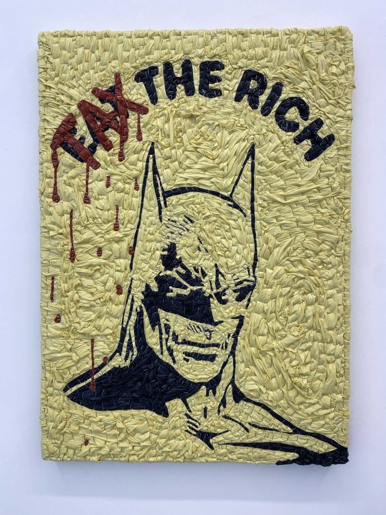 A composition made out of garbage bags stapled to a board showing a portrait of Batman above which is the slogan Eat the Rich in black but with the word "Tax" written over "Eat" in red and dripping like blood.