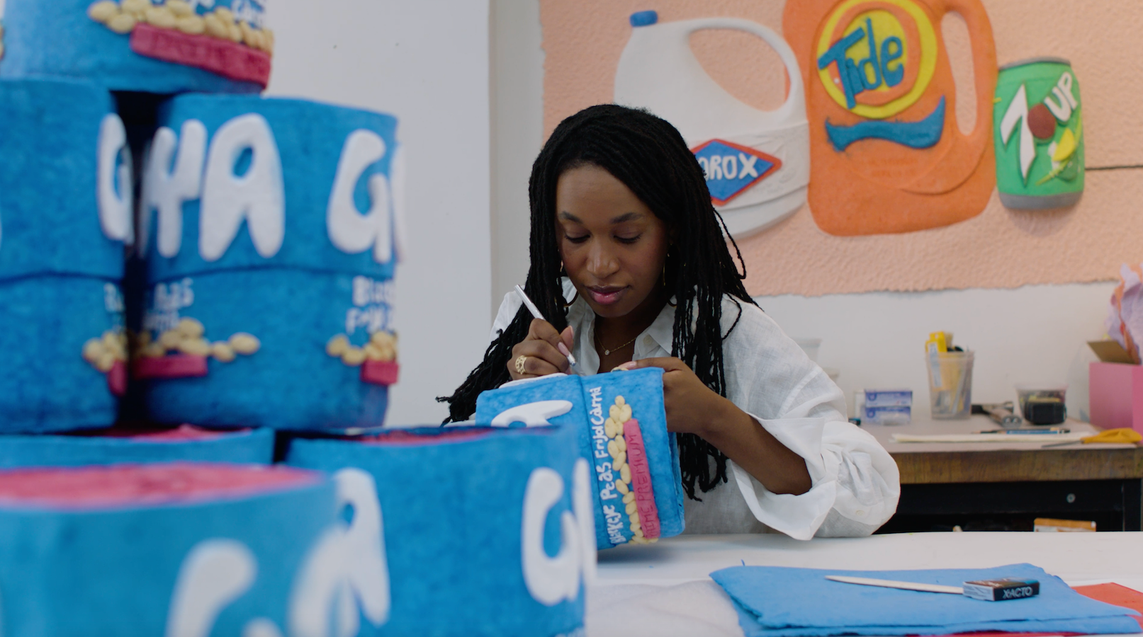 Tschabalala Self making her cast paper sculptures for "Bodega Run." A young Black woman, she is sitting behind a table on which there is a stack of blue cast paper sculptures of Goya cans. She is painting one. A cast paper still life of Clorox, Tide, and 7-Up is behind her.