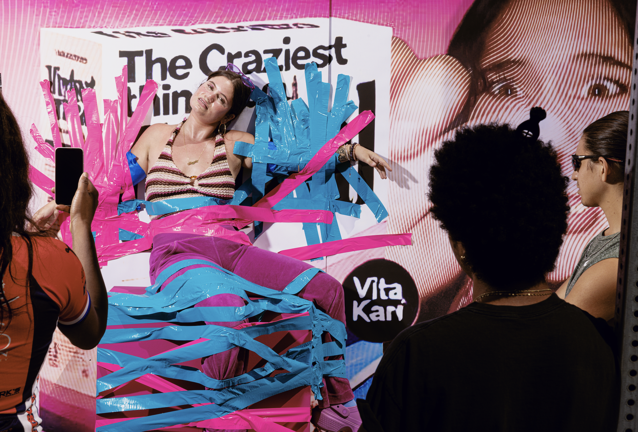 A photograph of Vita Kari affixed to a wall featuring an ad of themselves with blue and pink duct tape, as gawkers watch on.