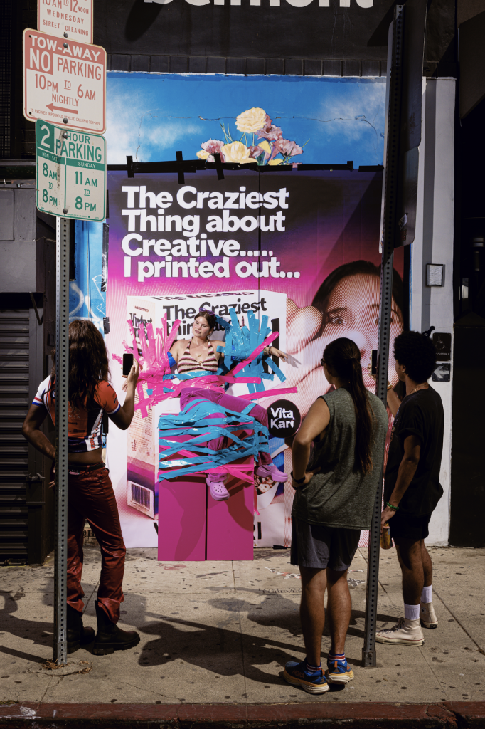 A photograph of a staged street scene in a studio, where onlookers gape at a person taped to a wall with blue and pink duct tape.