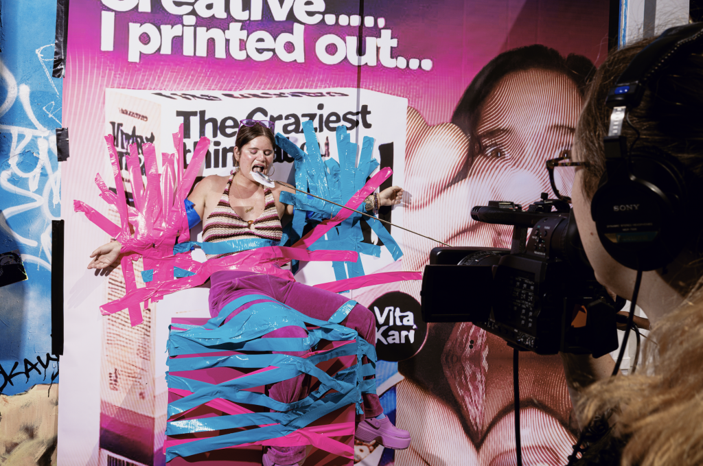 A photograph of a person affixed to a wall featuring an ad of themselves with blue and pink ductape, while a camera films the scene