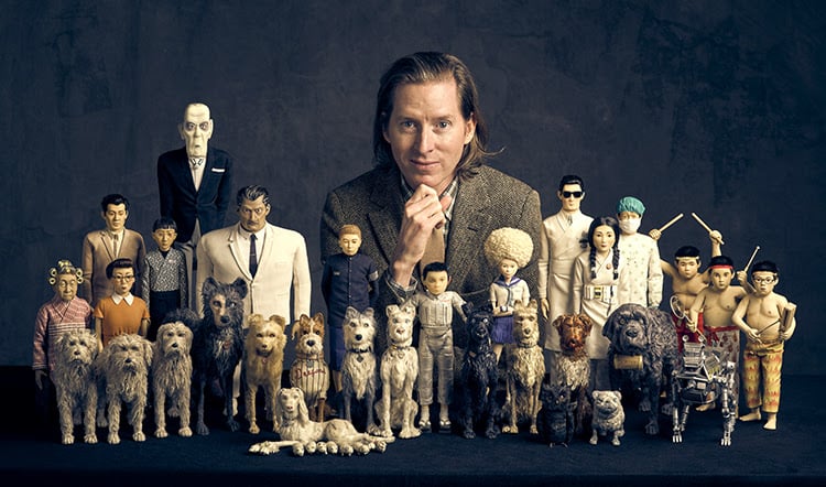 A photograph of filmmaker Wes Anderson seated with his chin on his hand behind a table of puppets that have featured in his films, amongst a flat dark-grey background.