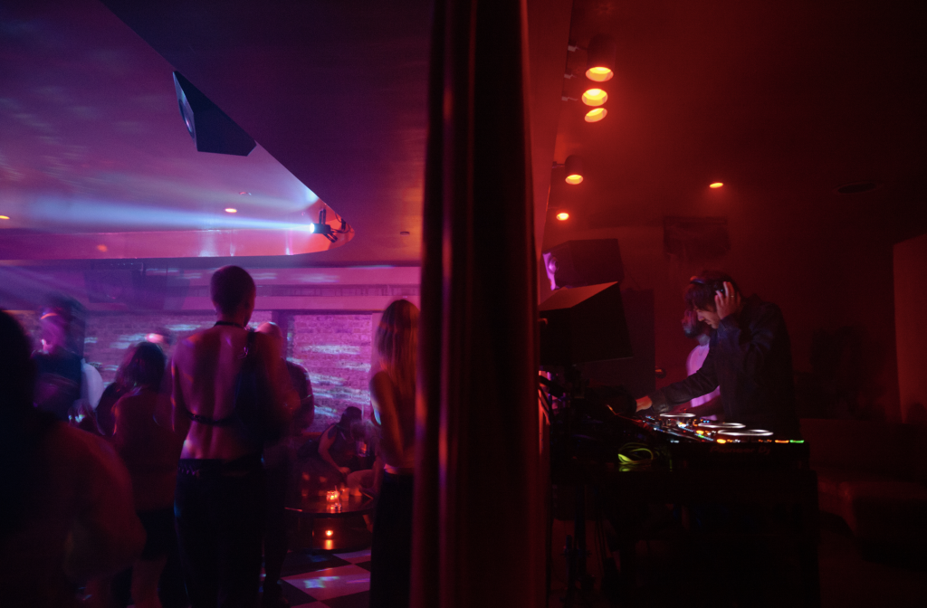 A photo of a moody party scene, with a purple-lit dance floor to the left and a red-tinged DJ booth on the other side of a divider, to the right.