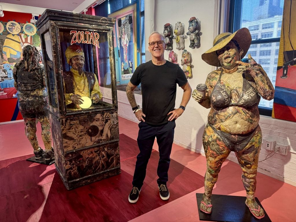 Will Kurtz with his Coney Island sculptures at Spring Break Art Show New York 2024. An older white man stands between a sculpture of a Zoltar fortune telling machine and a large woman in a bikini, both covered in newsprint.