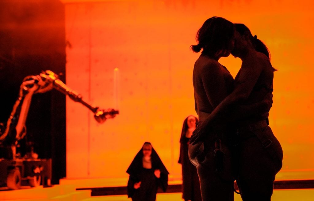 a glowing image of an opera dance art world with women embracing and a mechanical arm in the background