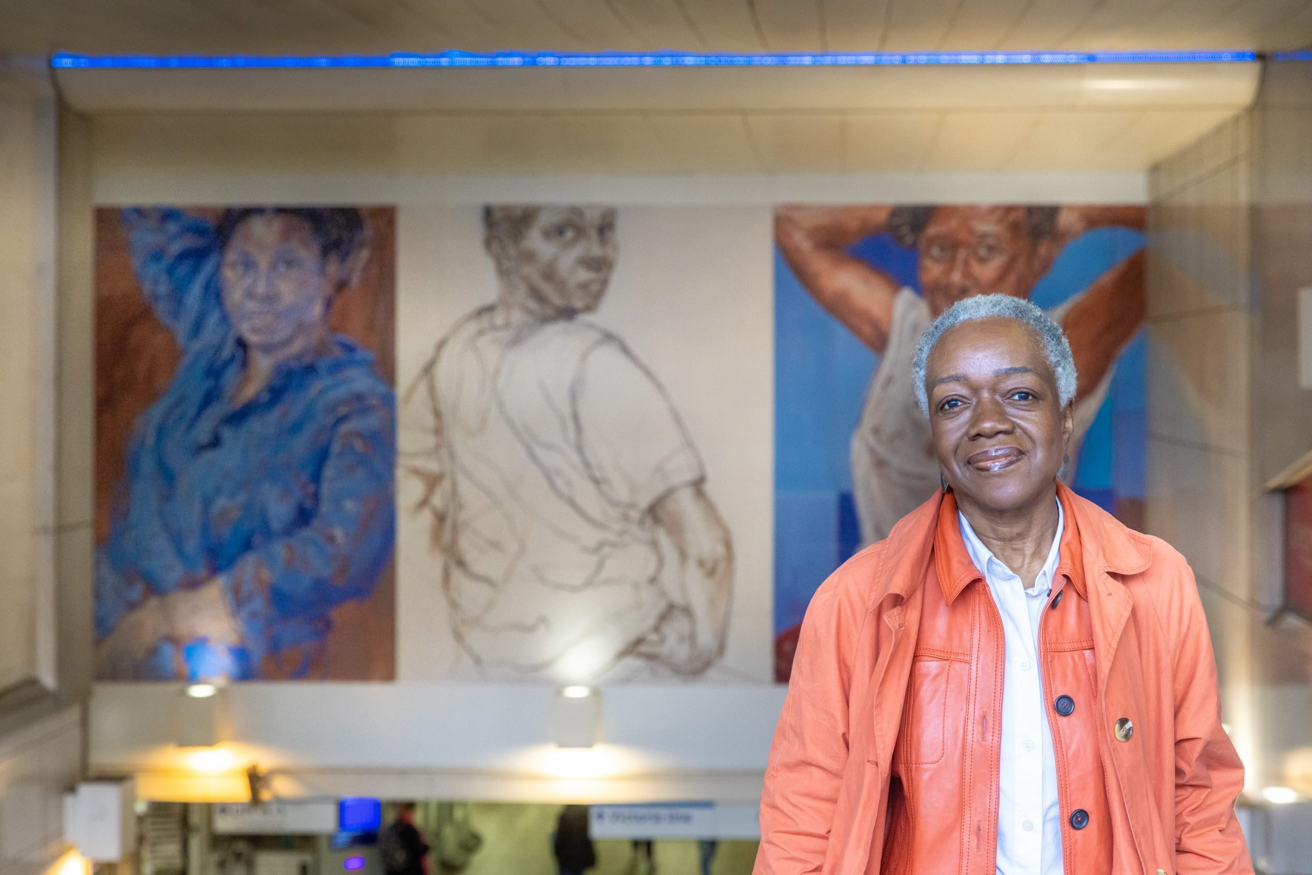 a woman with short white hair wearing an orange jacket stands in front of a mural depicting 3 women