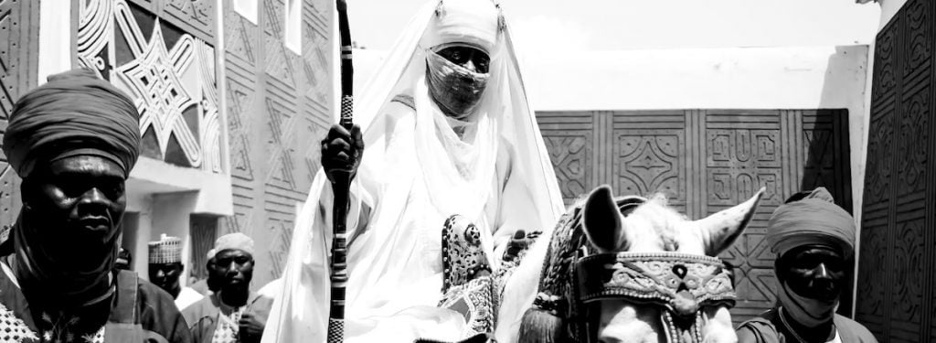 A black-and-white photo showing a group of men dressed in traditional African attire, with the central figure, possibly a ruler, riding a horse and holding a staff. The scene captures a moment of cultural importance, with intricate patterns on the surrounding structures.