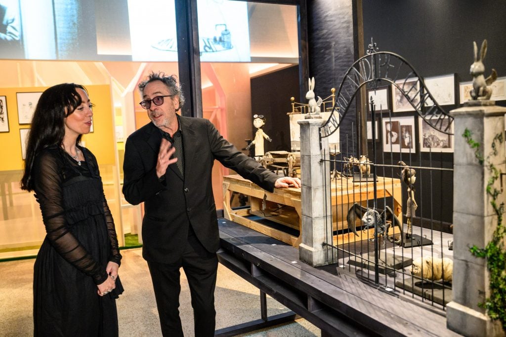 A man in black suit and sunglasses talks to a woman on his right hand side, his left hand touches models on display