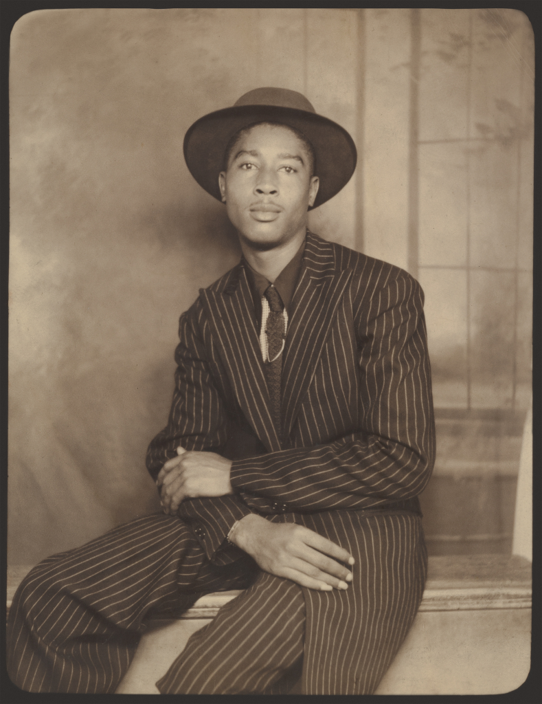 A black and white photo of a black man dressed in a striped suit and brimmed hat posing for a portrait. This man would be known as a dandy for his immaculate sense of style.