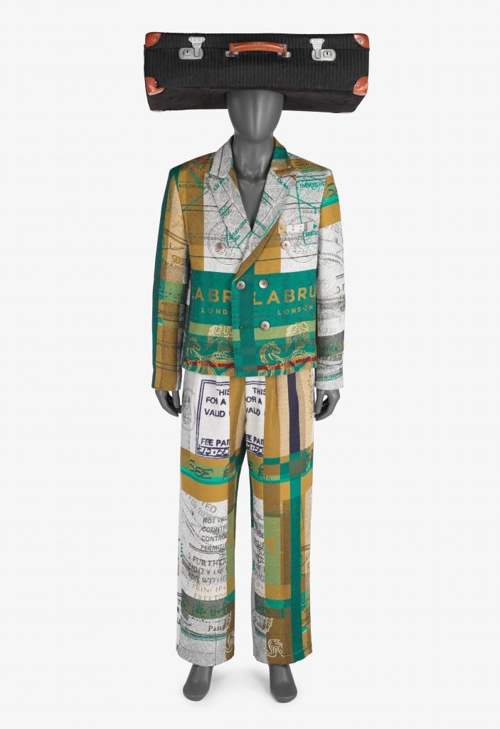 a colorful suit is draped on a mannequin. The design is reminiscent of passport pages and visa stamps. The mannequin is wearing a hat shaped like a suitcase