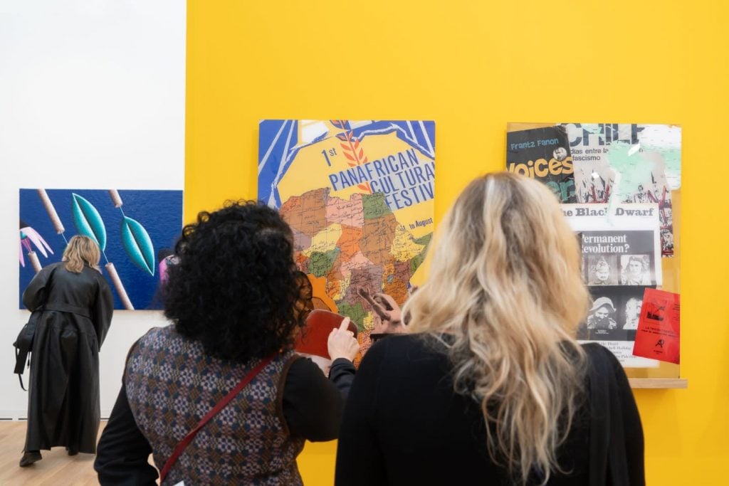 Two women examine vibrant artworks and posters, including a Pan-African Cultural Festival piece, in a brightly lit gallery.