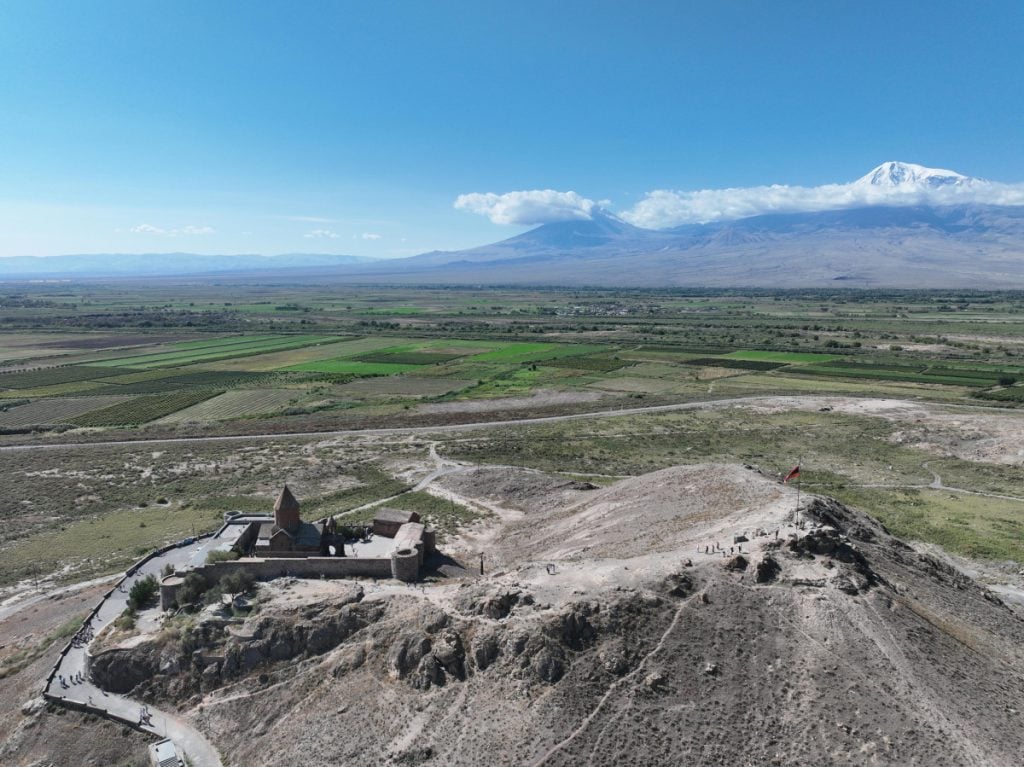 an aerial view looking over the plain with a white peaked mountain in the background and a monastery down below