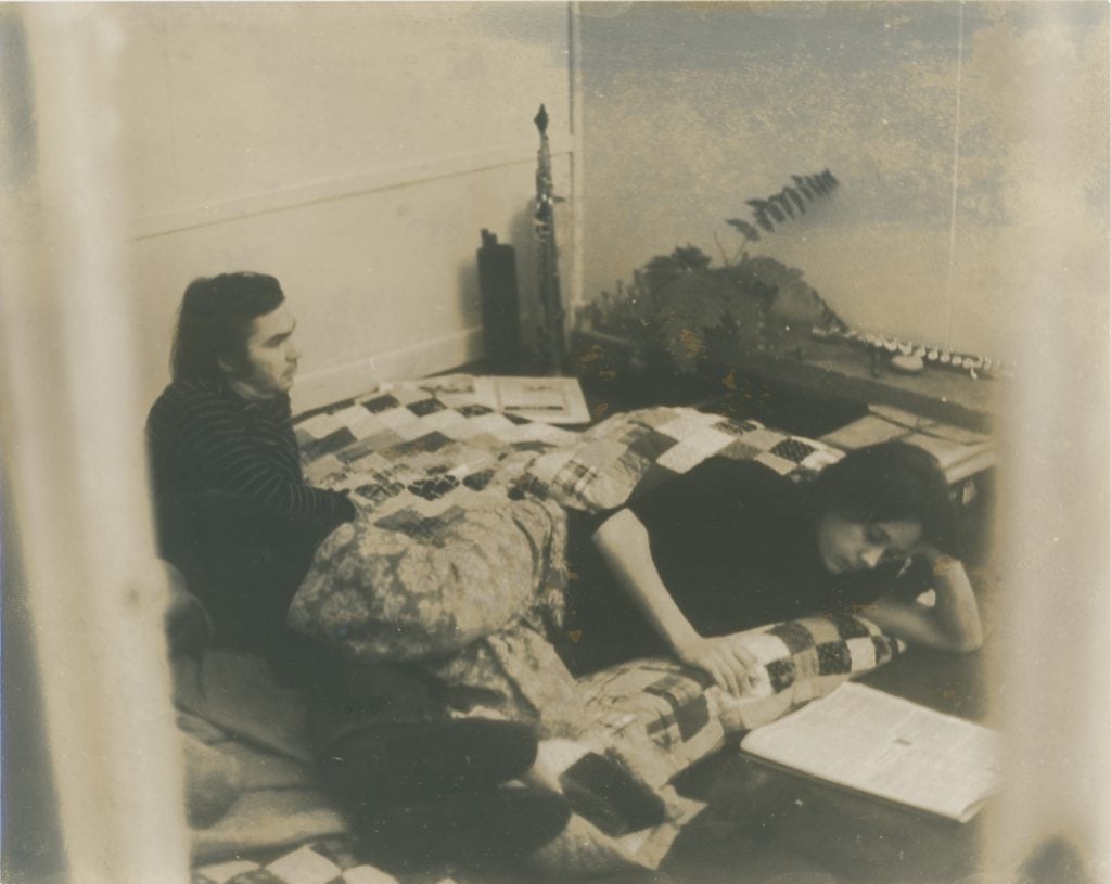 Black and white photo of Richard “Dickie” Landry and Tina Girouard at 10 Chatham Square in New York (ca. 1970s). The young white couple is lying on a mattress covere with a patchwork quilt. There are a few plants and artworks against the wall of the minimally furnished room.