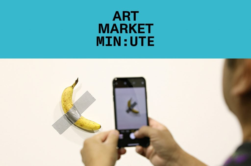 A person photographs a banana duct-taped to a wall as part of an art installation, under a banner that reads 