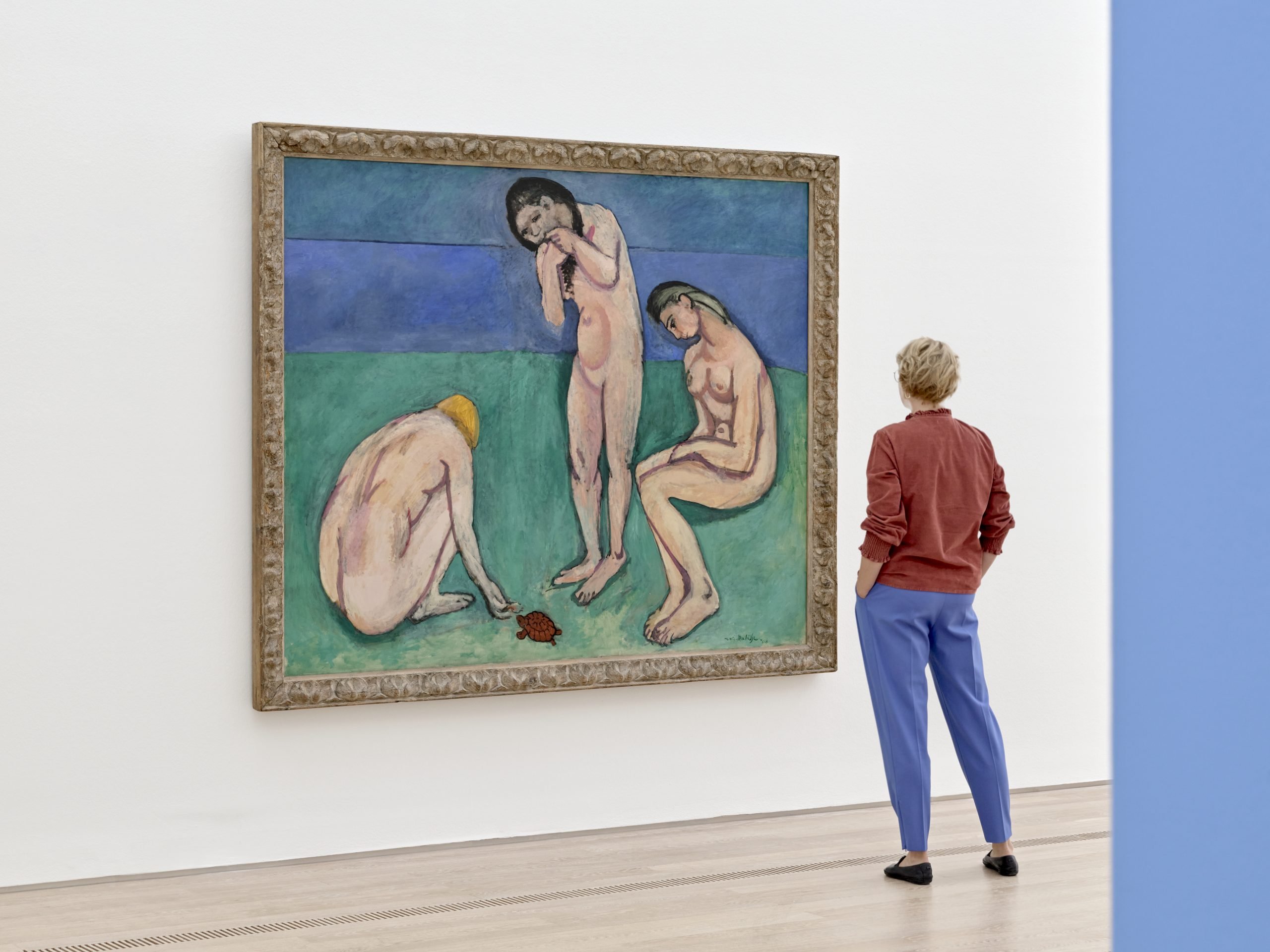 An exhibition goers enjoys the view of a large painting depicting three figures in nude against a backdrop of green and blue