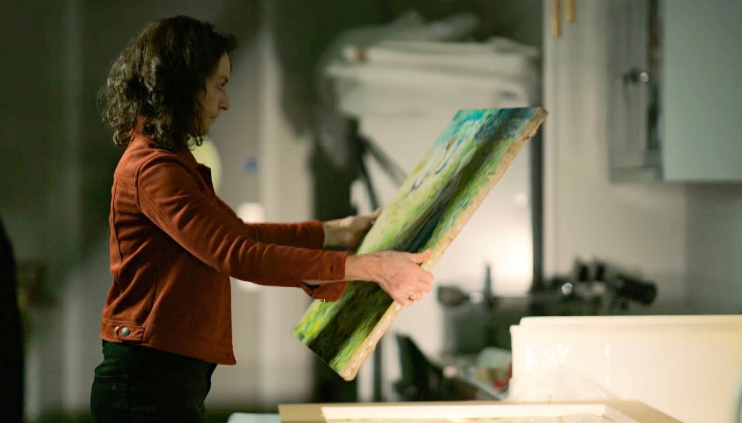 a woman holds up a painting in a dimly lit room