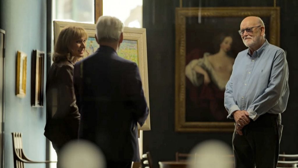 two people are seen from the back as they address a third man who stands in front of a painting