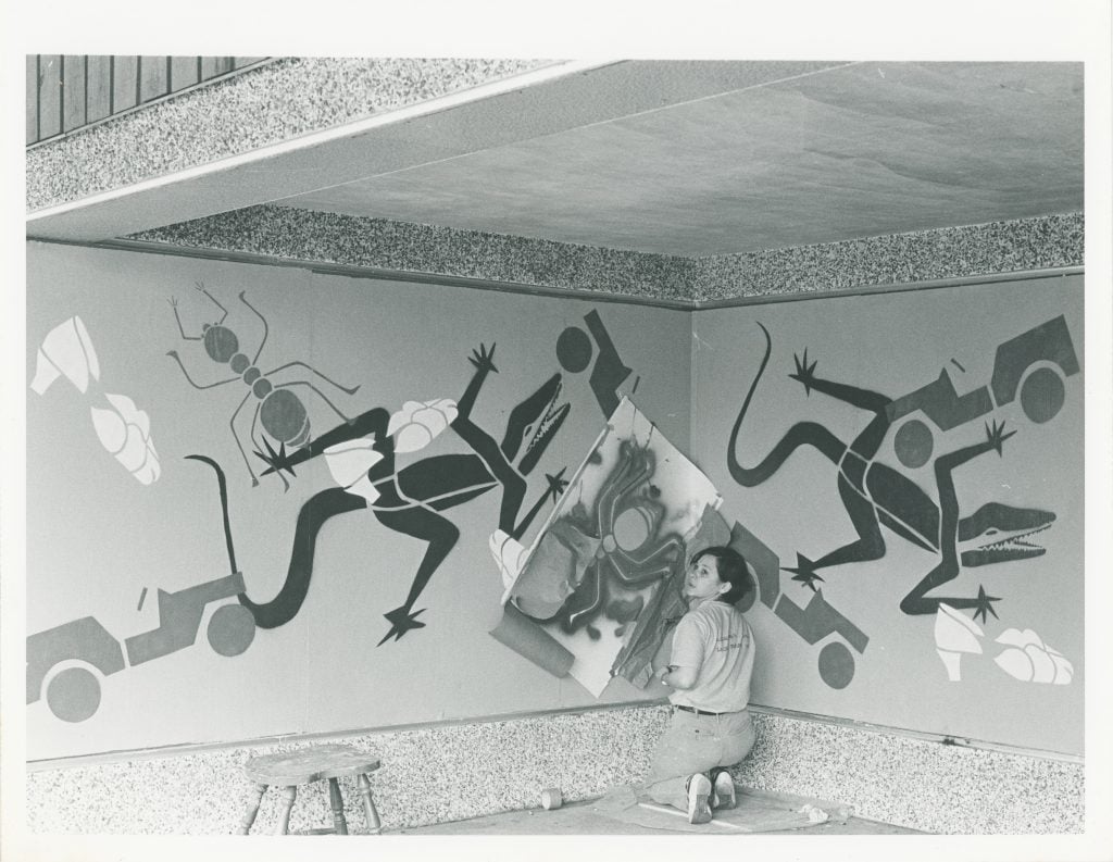 Black and white photo of Tina Girouard working on a stencil mural of stylized alligators, trucks, and shoes (ca. 1980s). 