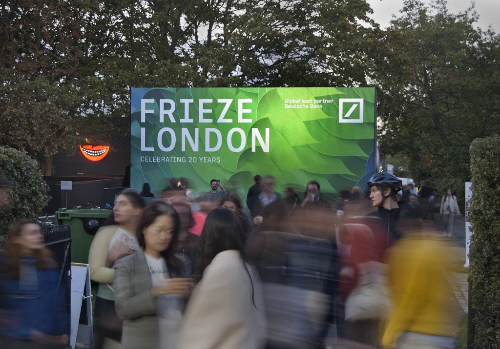 a crowd of people mills by a large green sign outside that says frieze london