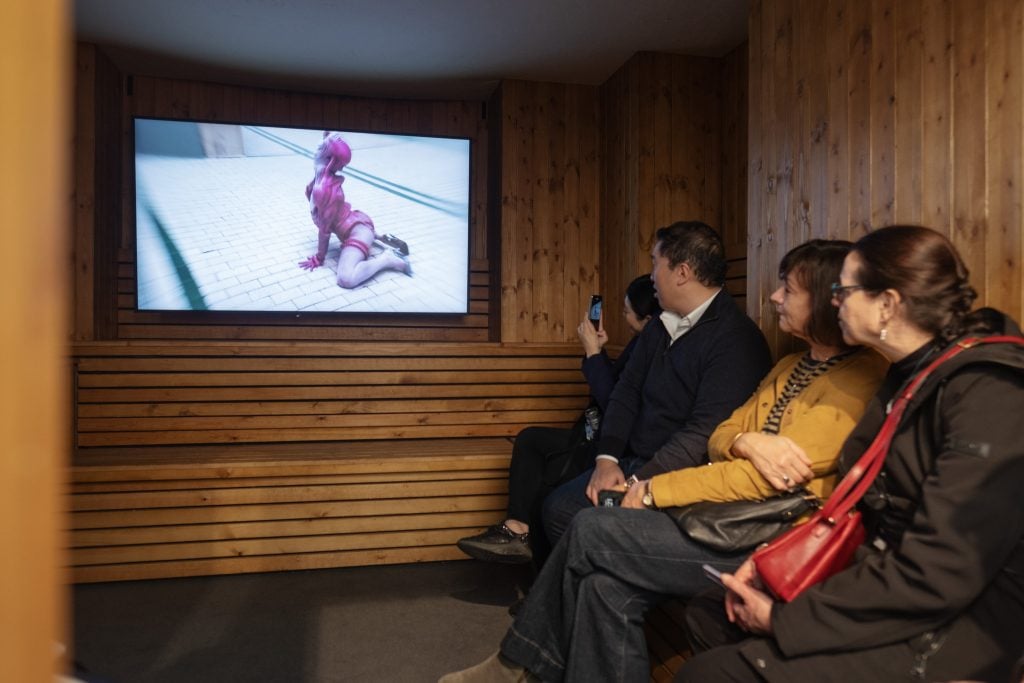 People watching a video installation inside a sauna-themed booth at an art fair.