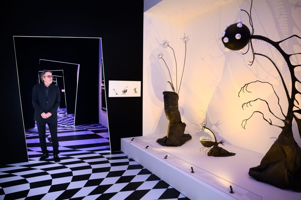 The director Tim Burton visits the exhibition 'The World of Tim Burton', he is looking at the monster looking sculptures placed in a room with black and white floor tiles and has many door frames