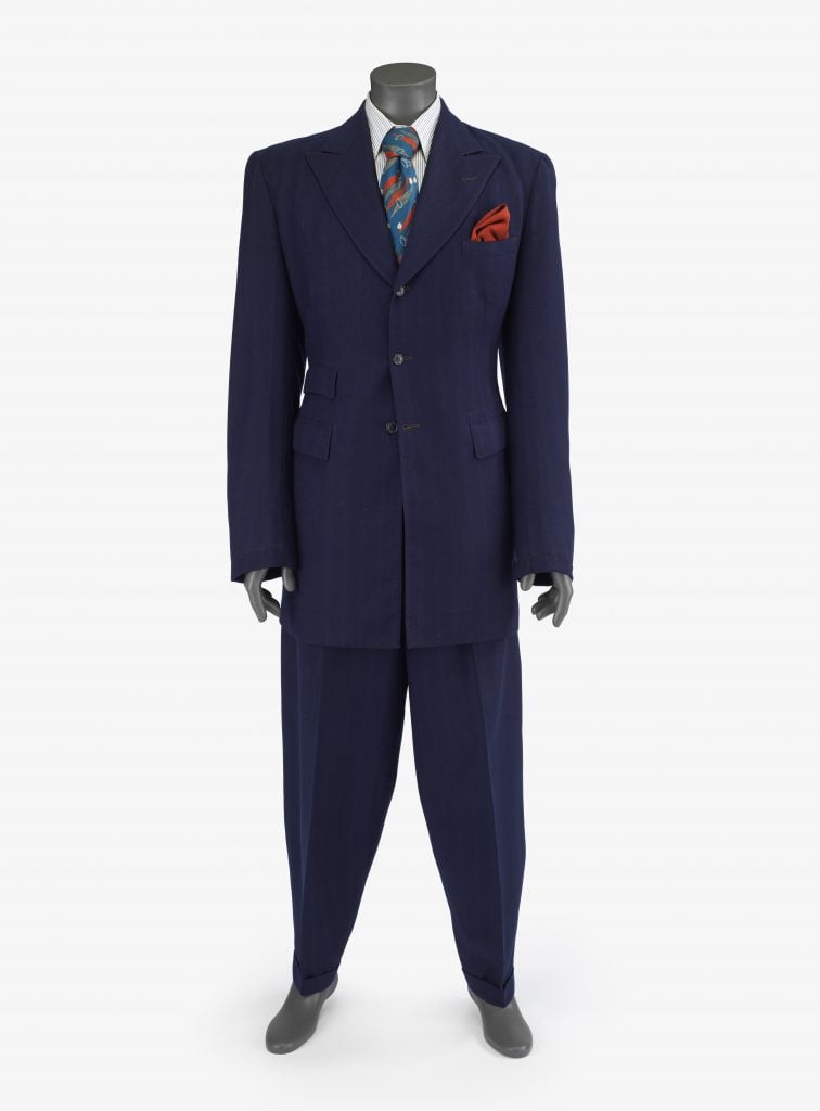 a zoot suit is draped on a mannequin, the suit is naxy striped twill, with dramatic proportions including an exaggeerated lapel long cuffs, and a cinched waist.