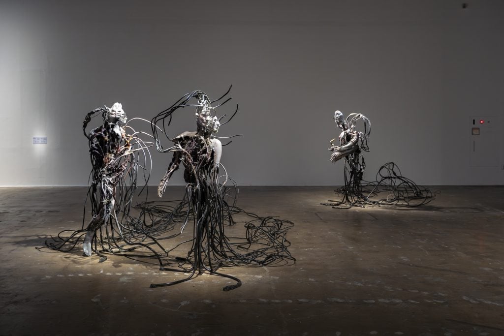 Three sculptures that look like humans but with lots of wires standing in a room