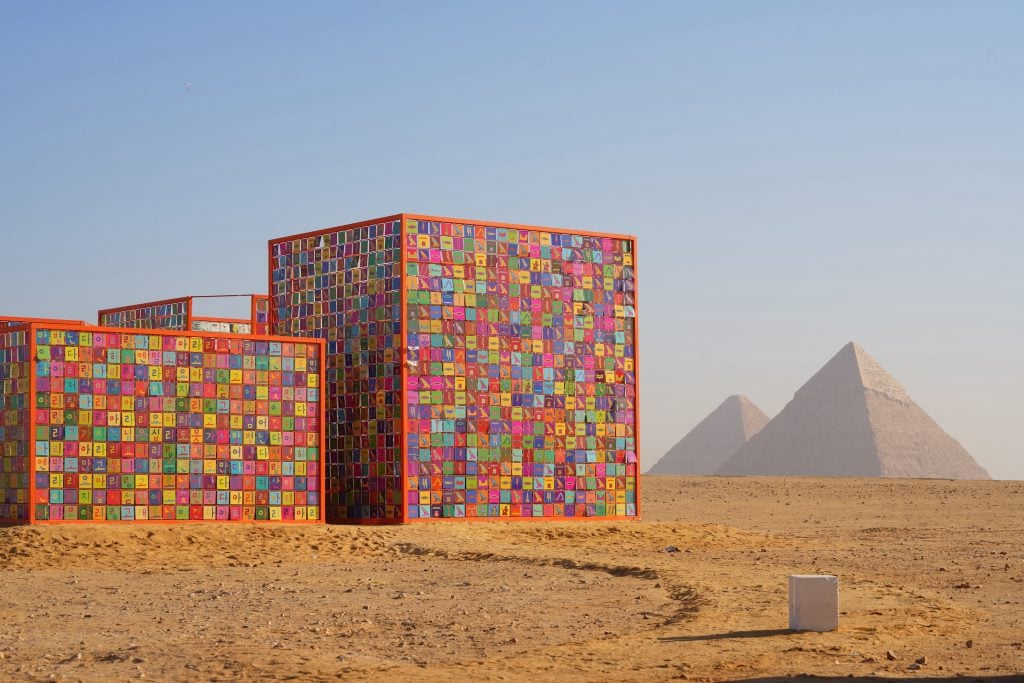 A colorful, cube-shaped installation covered in vibrant tiles stands in the desert with the Great Pyramids of Giza visible in the background under a clear blue sky.