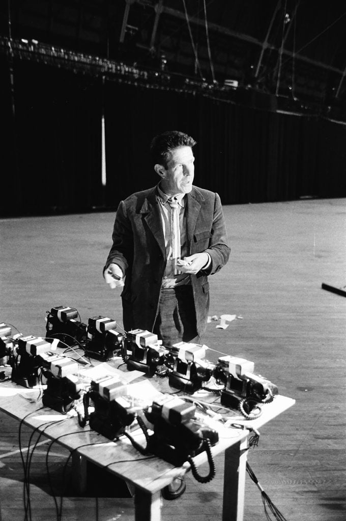 Composer John Cage in a tweed suit standing in front of a table arrayed with telephones