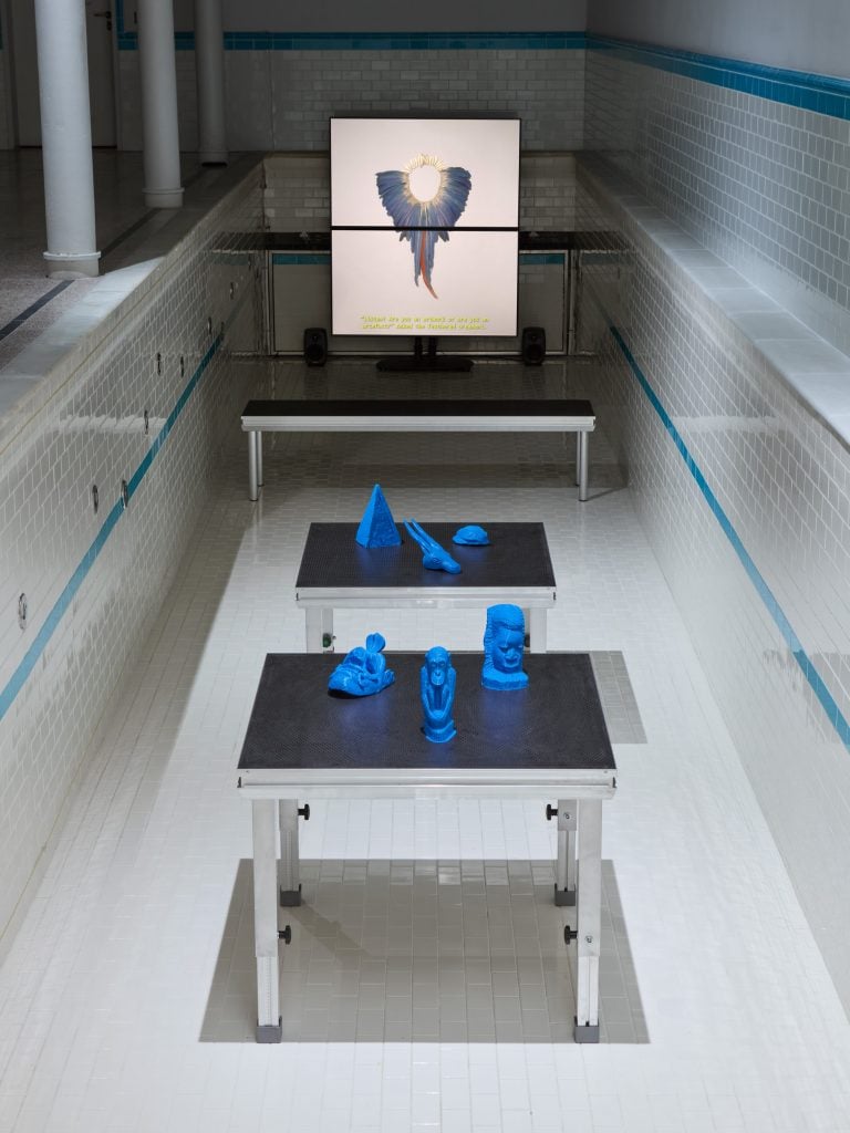 The image shows an art installation set inside an empty tiled swimming pool. Two metal tables in the foreground hold blue 3D-printed sculptures of various abstract shapes. A large screen is positioned at the far end of the pool, displaying an image of a circular object surrounded by what appears to be feathers, with text below. The pool's walls and floor are covered in white tiles with a blue stripe running horizontally along the top edge of the pool walls.