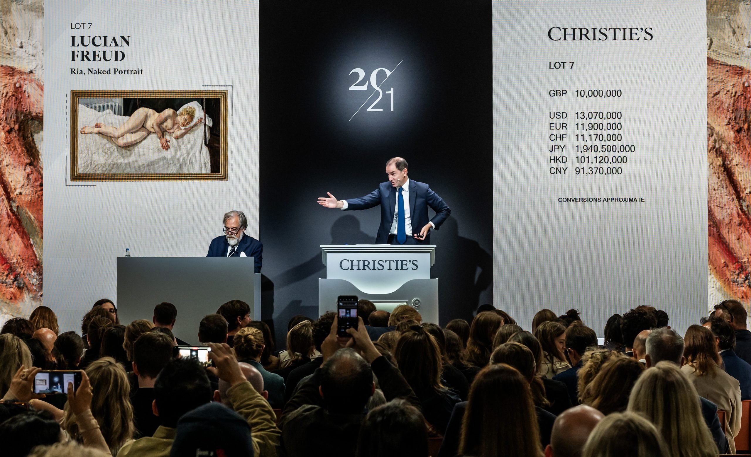 An auctioneer standing on the podium at Christie's selling a nude painting, against a backdrop with the sign of 20/21