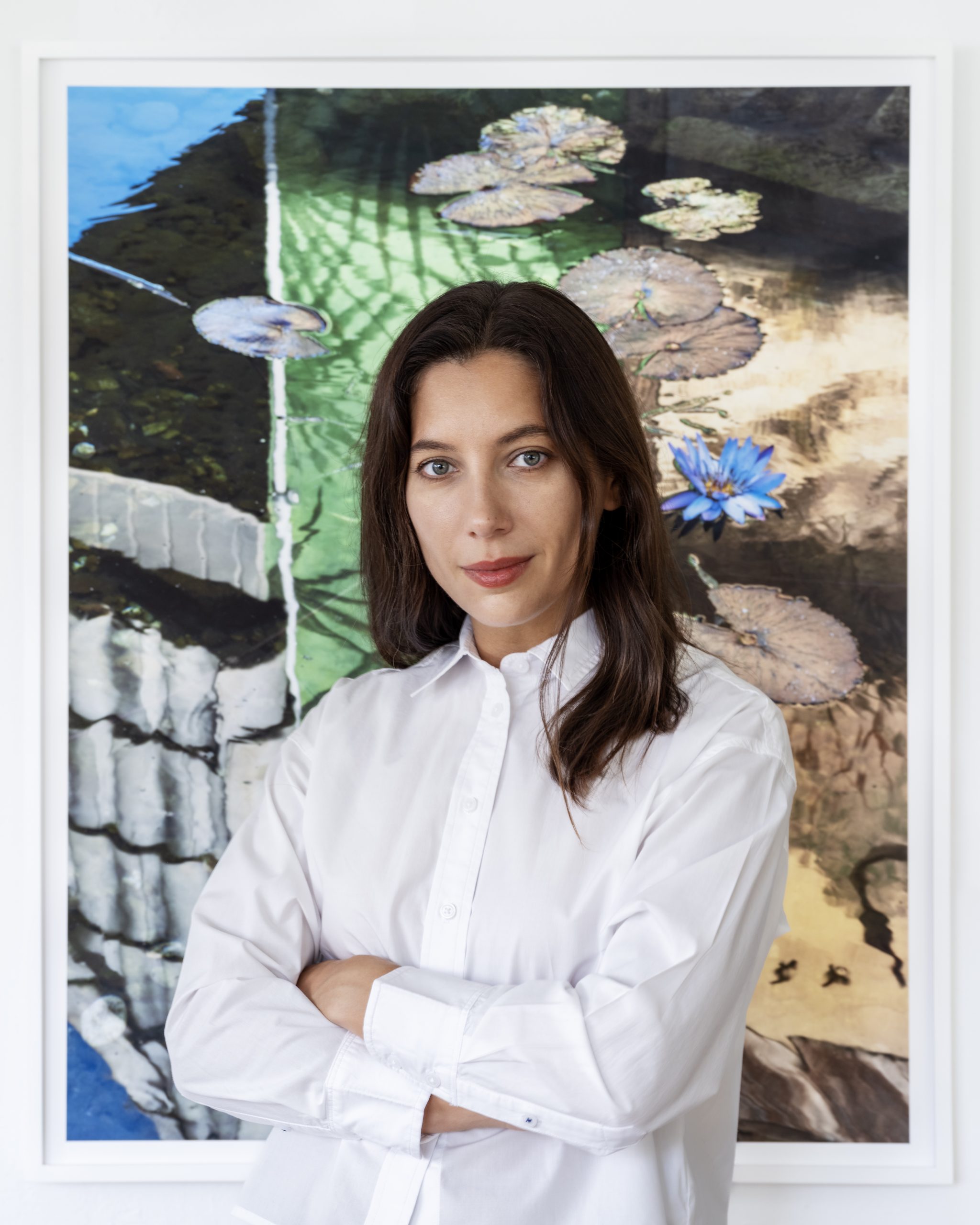 a white woman with brown hair stands in white button-down shirt front of a photograph with water