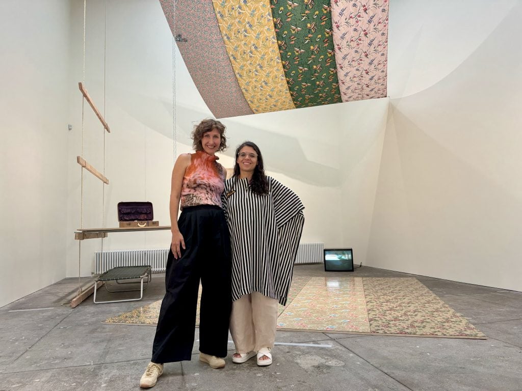 Two young, fashionably dressed curators, Andrea Andersson and Manuela Moscoso, stand in a gallery of work by Tina Girouard, including four lengths of floral silks suspended from the ceiling like a canopy.