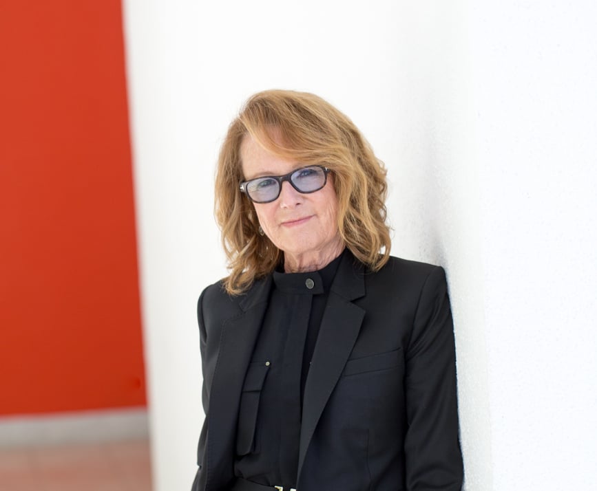 a portrait of a woman in glasses and a black suit with a black shirt leaning against a white wall