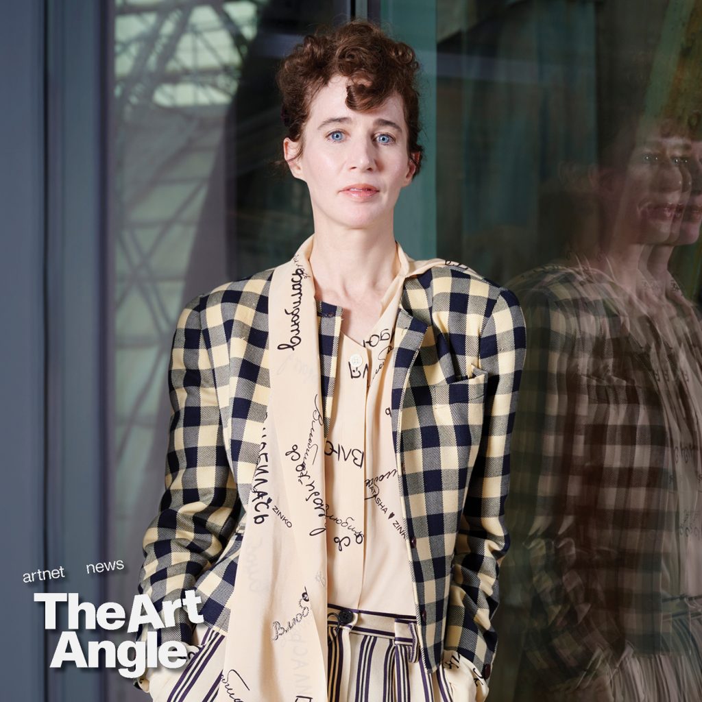 a fair-skinned woman with brown hair stands wearing a fashionable checkered blazer with a scarf around her neck