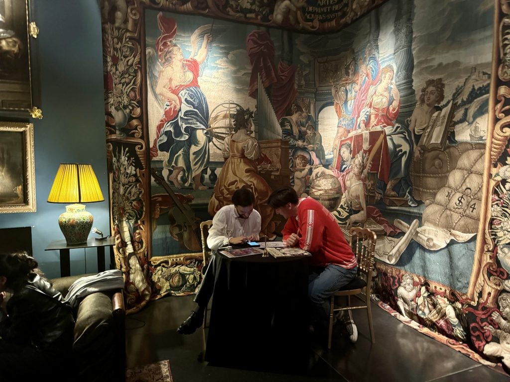 A dimly lit room holds tapestries and two people conducting an astrology reading