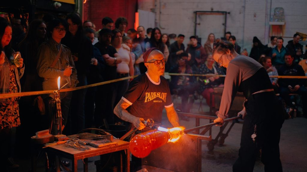 A glass blowing workshop at Brooklyn Glass. A crowd of onlookers stand behind caution tape as a man and a woman roll hot glass at a workstation.