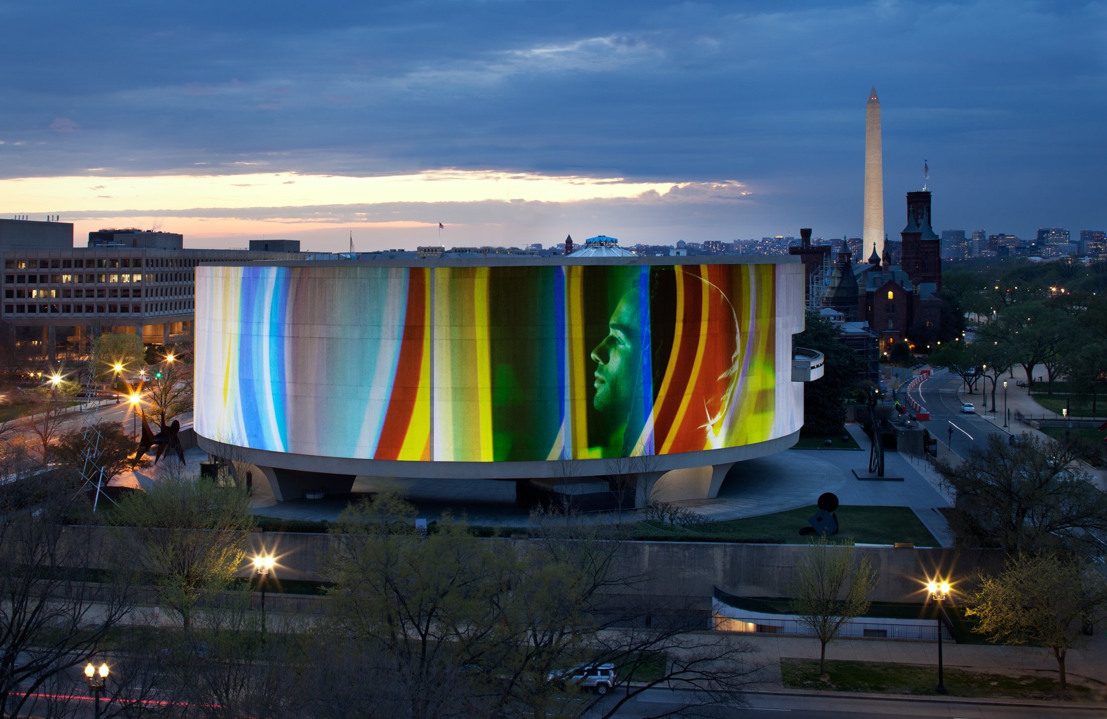 A cylindrical building displays a colorful, illuminated projection of a face at dusk, with the Washington Monument in the background.