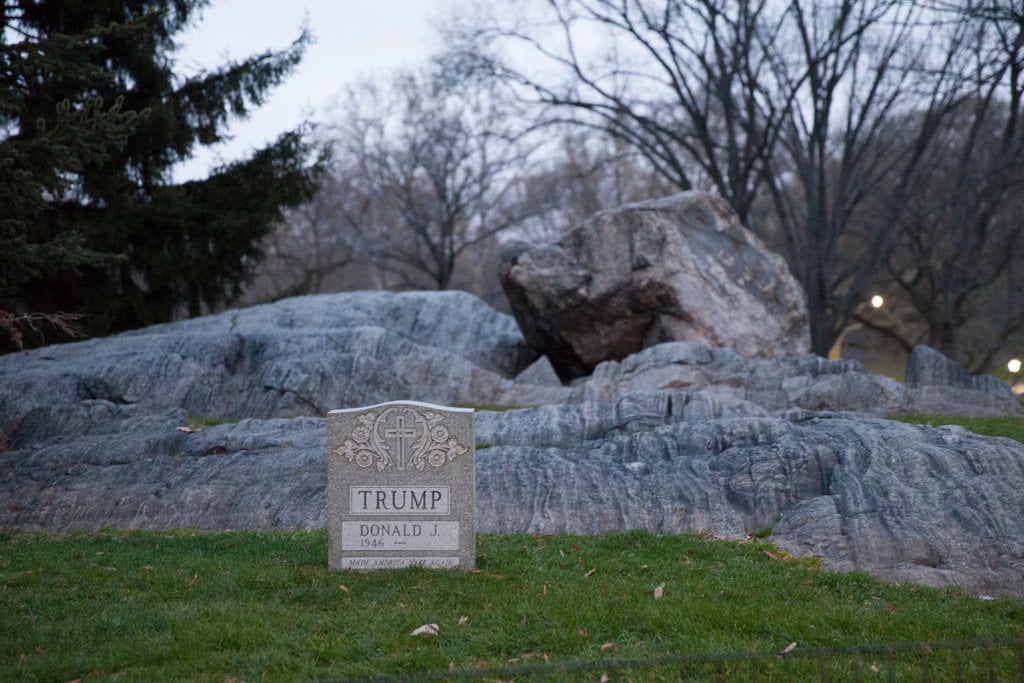 Brian Andrew Whiteley's Donald Trump tombstone, Legacy Stone, in Central Park's Sheep Meadow on Easter Sunday, March 2016. The carved gravestone has an ornamental cross, Trump's name, and the inscription "Made America Hate Again." It site on the grass in front of stone boulders, with several trees in the background. 