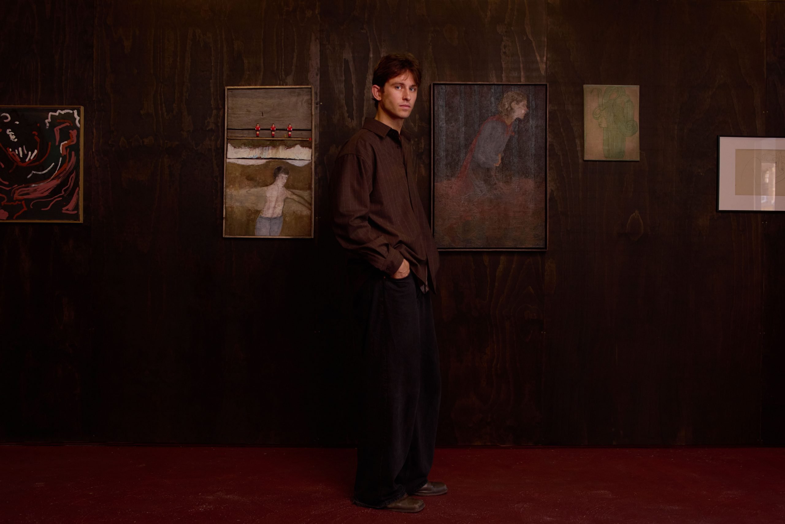 a man in brown stands in a very dark room surrounded by paintings