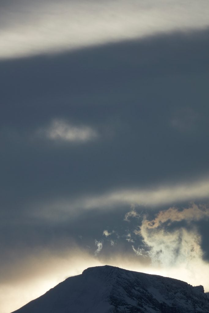 Catherine Opie, There's always Norway... (2024). The photo is a vertically oriented landscape, the top of a dark hill seen against a wispy cloud with a dark blue sky above. 