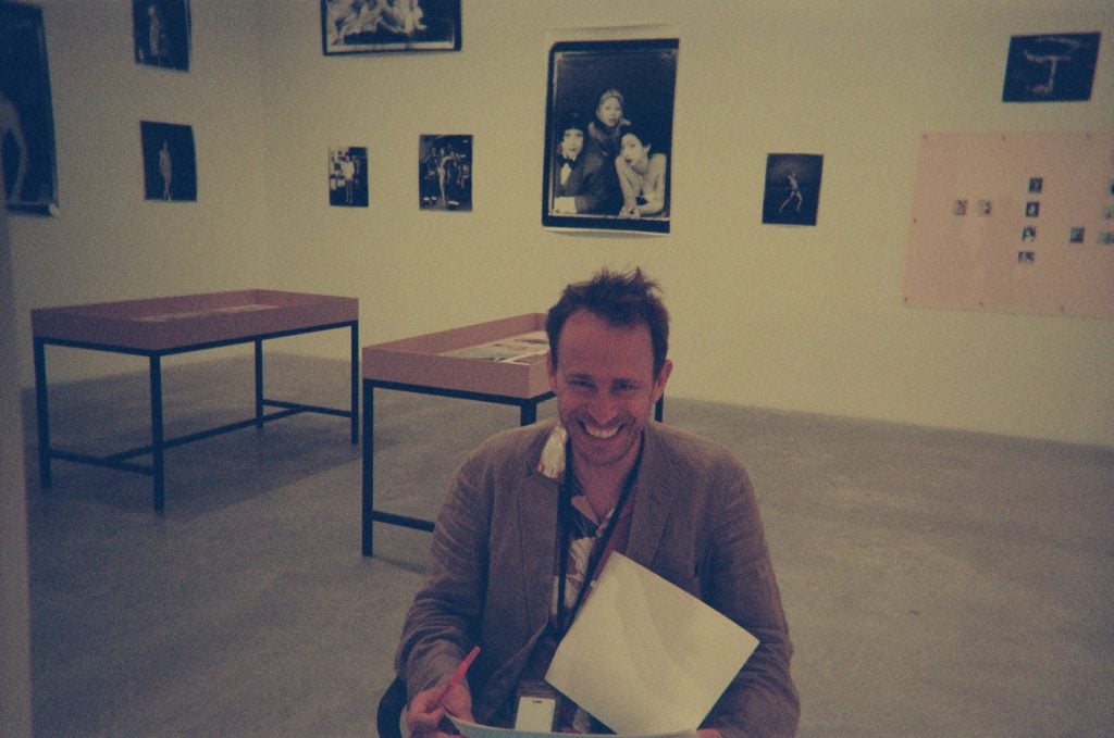 a white man sits inside a gallery room with photographs on the walls