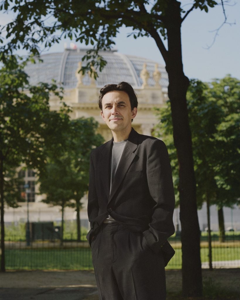 Man in a black suit stands beneath trees, grand building in the background.