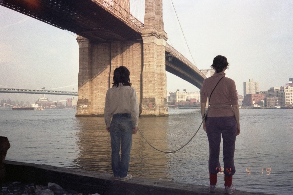 A man and a woman, tied together with a several foot long rope, look out over a river under a bridge