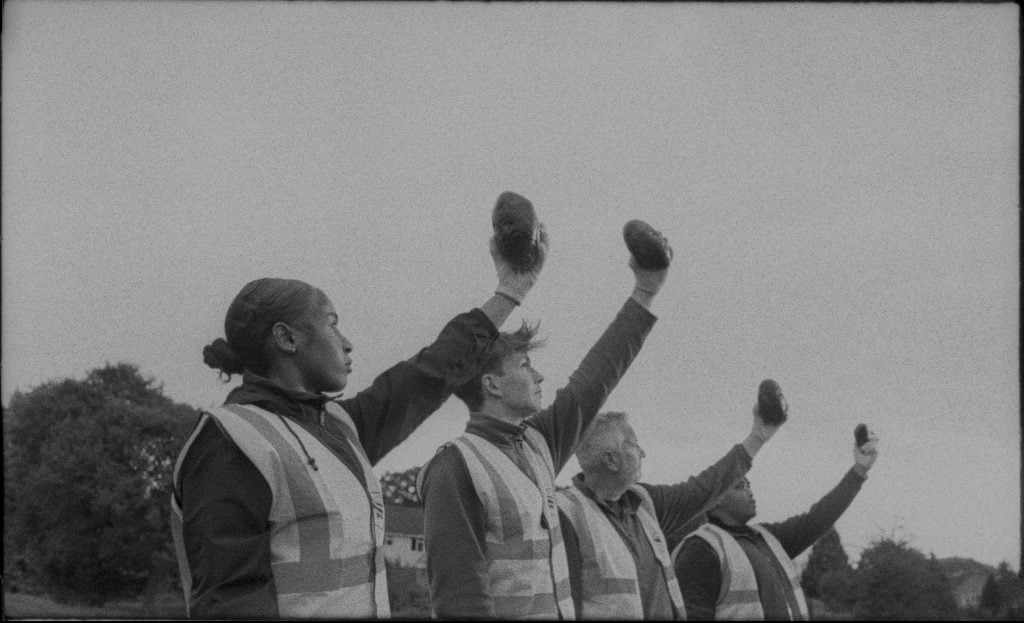 a black and white film still of 4 people standing in a line and holding up their left arm with a rock in their hand