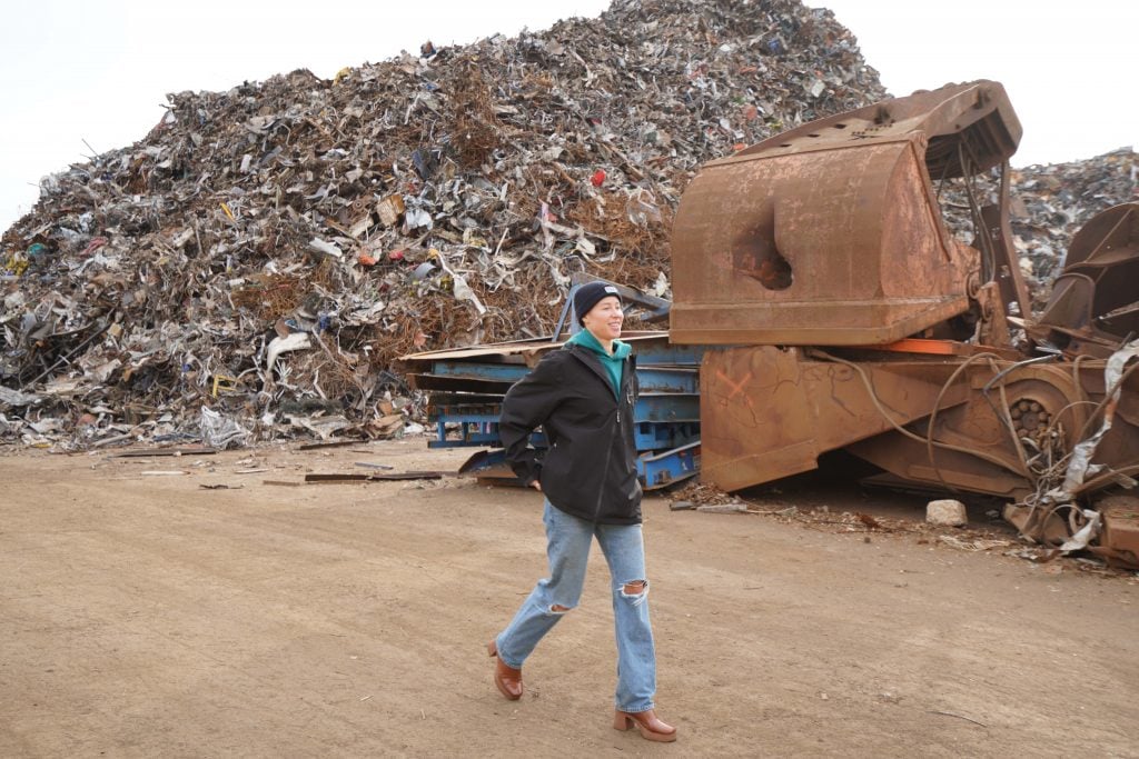 a woman with jean, jacket, and beanie walks through a giant scrap yard