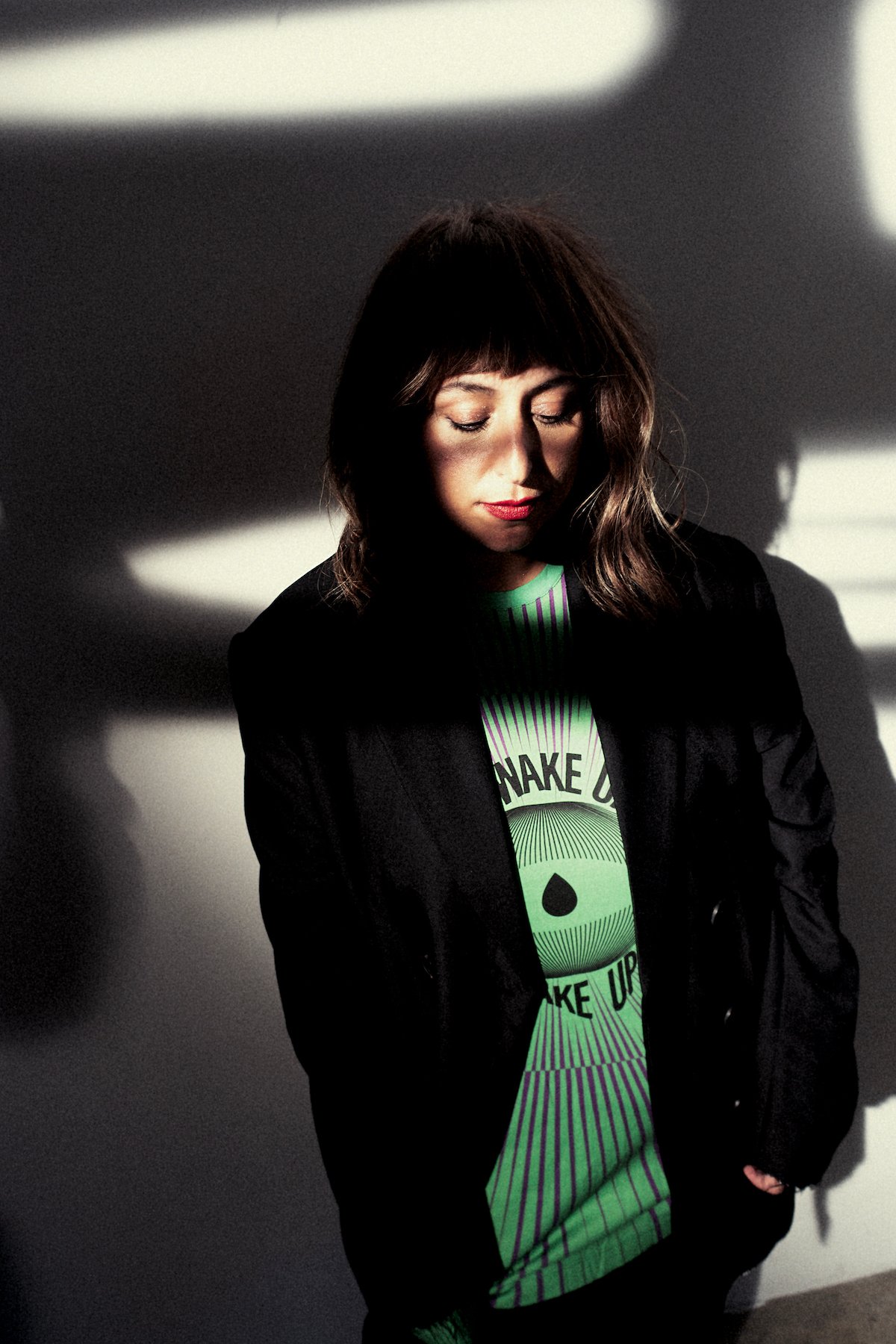 A woman in a green tshirt, black blazer and red lipstick stands in shadow